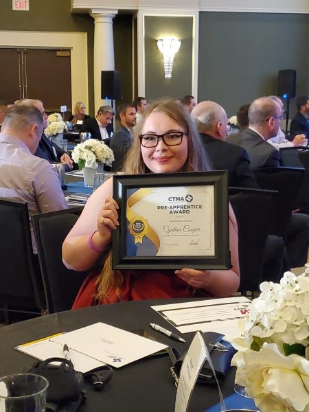 Pre-Apprentice EDM Operator Cynthia Cooper poses with her Pre-Apprentice Award while sitting down at her table at the CTMA AGM. The Award is a certificate in a black frame.
