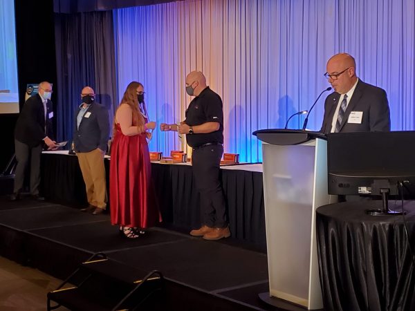 Cynthia Cooper walks across the stage at the CTMA AGM to accept her award. CTMA Director Robert Cattle is speaking into a microphone on the right side of the stage.