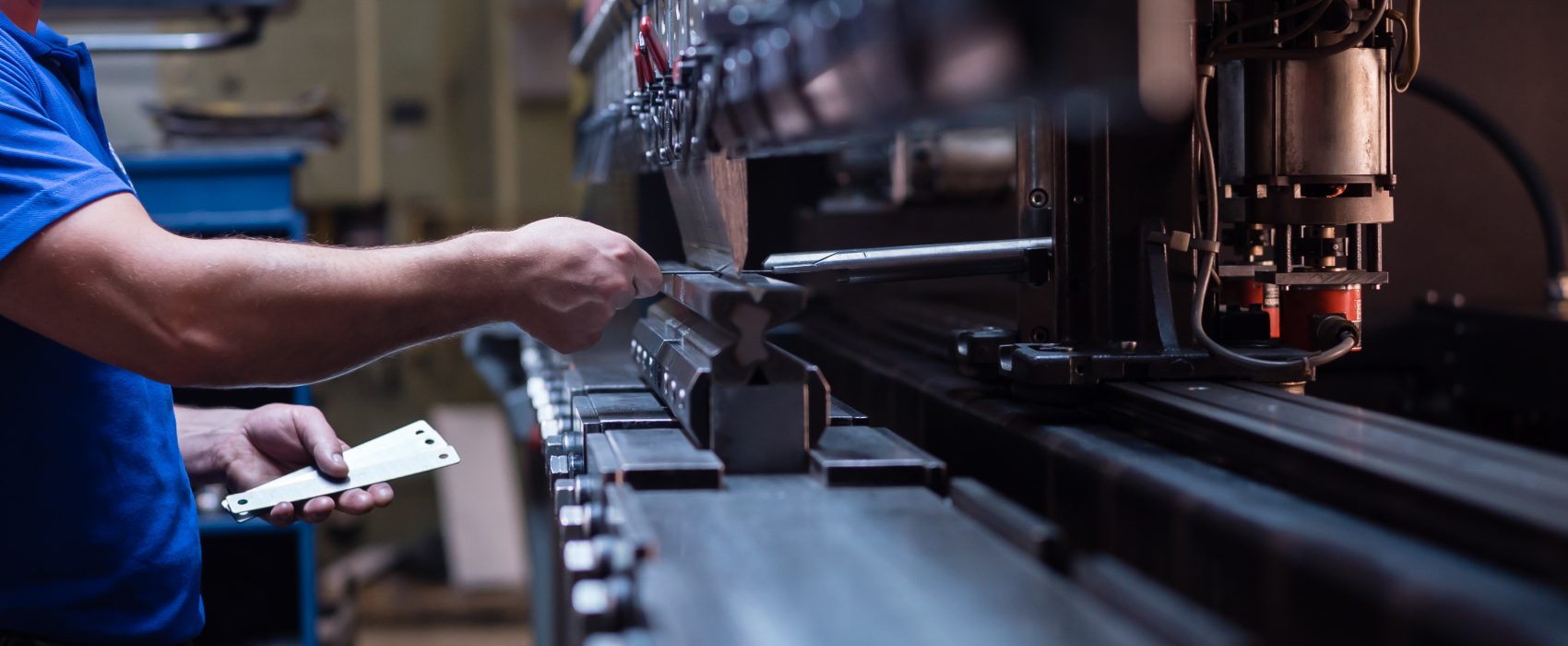 right side close-up of a worker holding a metal plate against a manufacturing machinery, in the right hand, and several other metal plates in his left hand